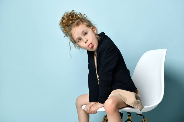 Naughty little girl dressed in business style wearing her curly hair up sitting in office chair leaning forward and lolling out, isolated on blue - Valokuva, kuva