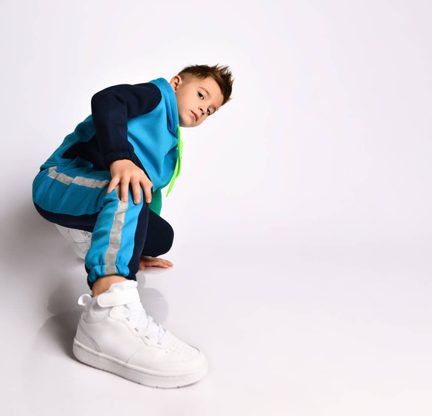 Little son in colorful tracksuit, sneakers. He performing exercises or kick by his leg, posing isolated on white background - Foto, Bild