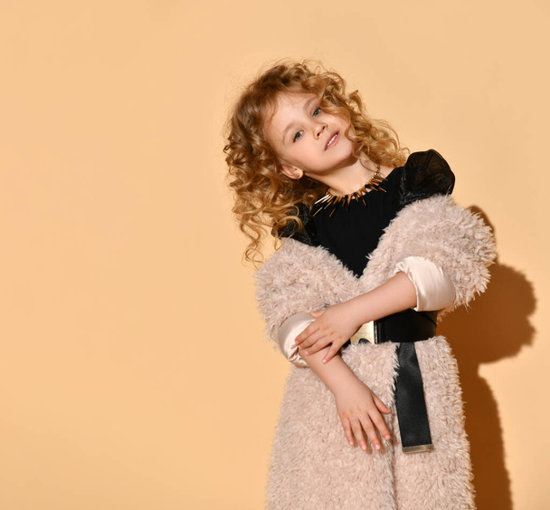 Pretty blonde curly model in necklace, black dress and faux fur coat with belt. She is smiling, posing against beige background - Zdjęcie, obraz
