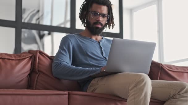 A serious young african american man wearing eyeglasses is using his silver laptop computer sitting in the living room at home - Záběry, video