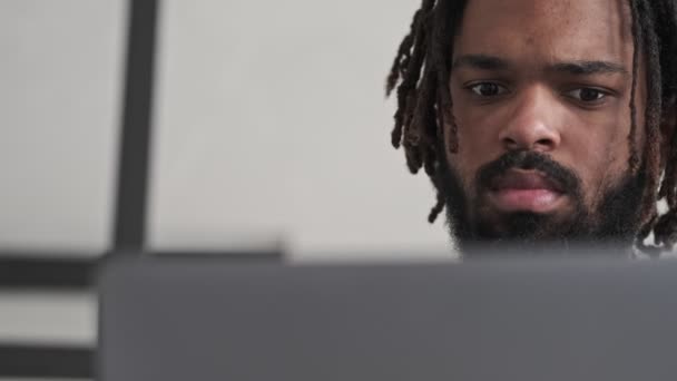 A close-up view of a serious african american man is using his silver laptop computer sitting in the living room at home - Video