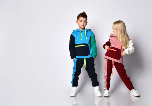 Little kids, boy and girl, in colorful tracksuits and sneakers. They posing isolated on white studio background. Hands in pockets - Φωτογραφία, εικόνα