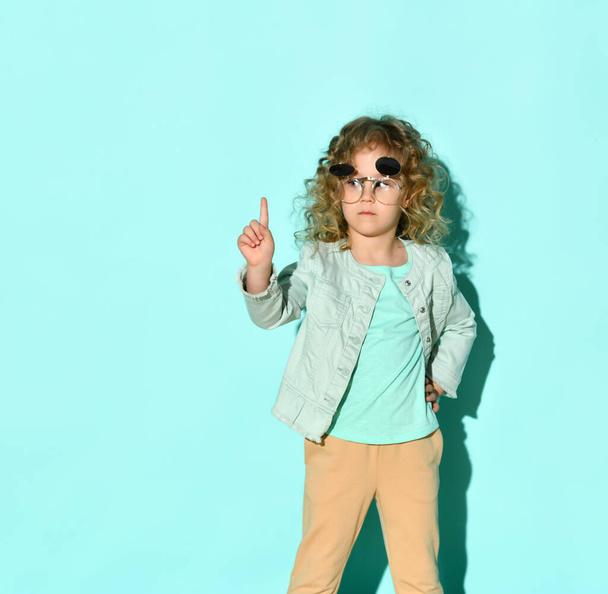 Serious little girl in jacket, blue t-shirt and beige jeans posing in flip-up glasses with her finger up asking to wait - Фото, изображение