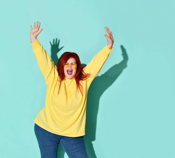 Red-haired voluptious young woman screaming with closed eyes and showing stop by both hands. Three quarter length portrait isolated on blue - 写真・画像