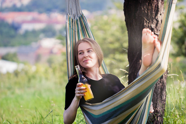 Una donna felice con i capelli biondi sta distendendosi e sorridendo in un hammock all'aperto con una bottiglia della bevanda gialla rinfrescante su un giorno d'estate soleggiato. - Foto, immagini
