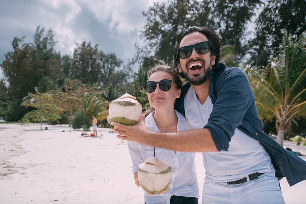 Пара влюбленных веселятся гуляя по тропическому пляжу с коктейлями. Парень и девушка на свежем кокосовом фоне из тропических растений и пальм. Молодожены наслаждаются белоснежной Лагуной
 - Фото, изображение