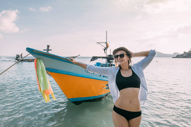 Молодая женщина на тропическом пляже возле рыбацкого судна. Веселая девушка в бикини и шляпе позирует на фоне моря и цветной рыбацкой лодки
. - Фото, изображение