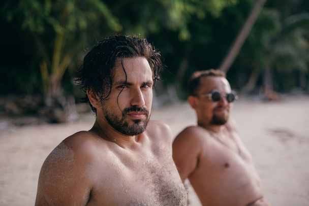 Yakışıklı adamlar okyanusun kenarında kumların üzerinde oturuyorlar. Gençler - tropik bir plajda çıplak bir gövdeyle güneşleniyorlar.. - Fotoğraf, Görsel