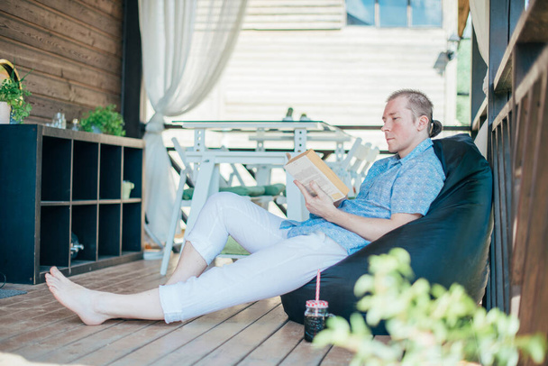Un hombre lee un libro en el porche. Joven chico guapo se encuentra en una otomana con un libro en la terraza de una casa de campo en un día soleado - Foto, imagen