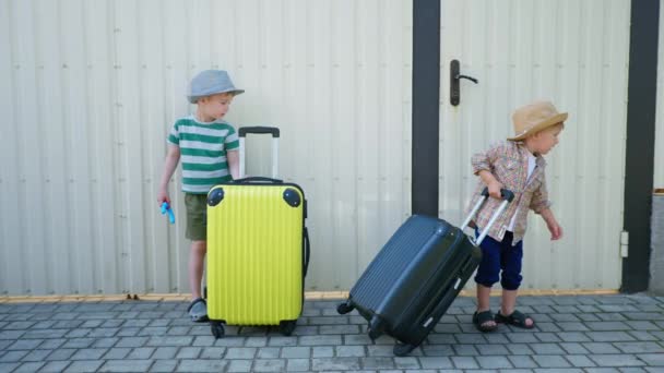 viaggio per bambini, piccoli bambini maschi attraenti in cappelli da sole si divertono vicino a valigie durante le vacanze estive sullo sfondo di recinzione bianca - Filmati, video