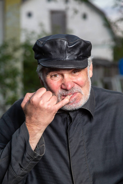 Un vieil homme vêtu de vieux vêtements noirs et coiffé d'une casquette en cuir regarde le spectateur et prend sa dent avec son petit doigt. Fond de campagne flou. Orientation verticale.  - Photo, image