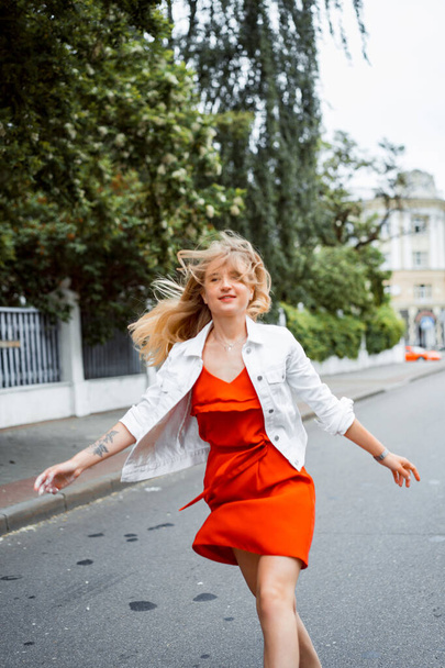 gyönyörű fiatal nő piros ruhában és fehér farmerdzsekiben, boldog mosollyal járja körbe a várost. Nyár. - Fotó, kép