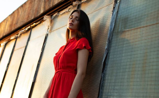 Profile portrait of a sensual young woman in red dress, with makeup, has got closed eyes, dreaming, posing on roof, near old black wall. - Photo, Image