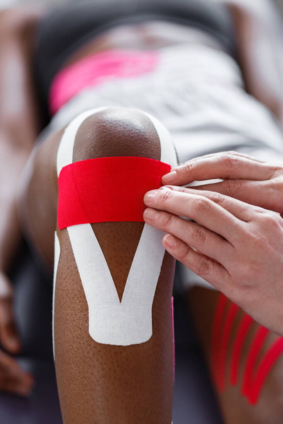 Physiotherapist applying kinesiology tape to patient knee.Therapist treating young female African American athlete. Kinesiology taping. Post traumatic rehabilitation,sport physical therapy - Zdjęcie, obraz