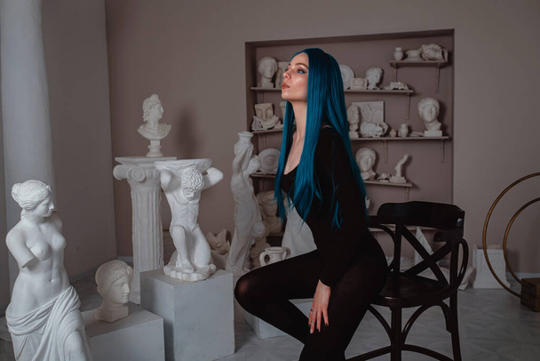 Beautiful young girl with blue hair in skinny black at the working place in the old atmospheric studio. sitter model, muse and inspirer concept - Photo, Image