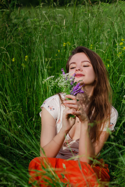 Young brunette girl in white top and red skirt sitting in green field grass in countryside smelling small bouquet of field flowers in hands with closed eyes and enjoy nature and rural life.  - Foto, afbeelding