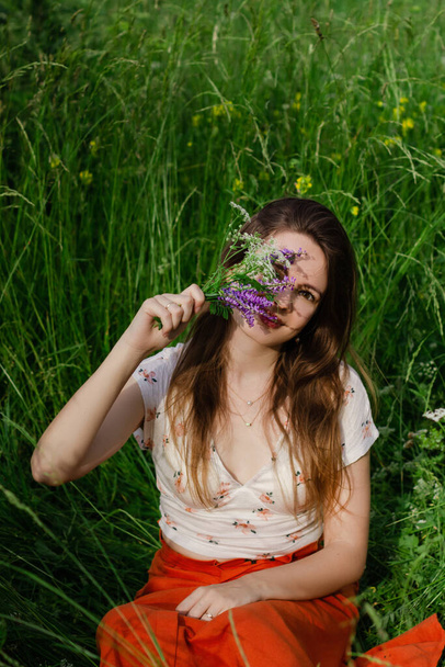 Young brunette girl in white top and red skirt sitting in green field grass in countryside smelling small bouquet of field flowers in hands with closed eyes and enjoy nature and rural life.  - Φωτογραφία, εικόνα