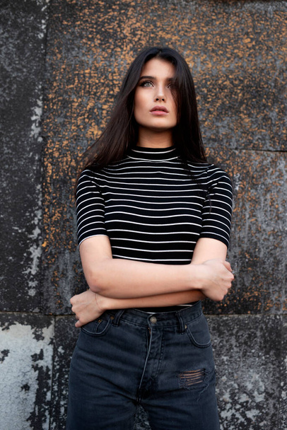 Single girl with straight brown hair and makeup, wearing black casual clothes, holding her hands crossed, on the black background of the old wall. - Zdjęcie, obraz