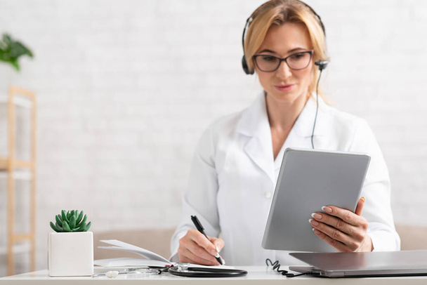 List of symptoms. Woman doctor with headphones holding tablet in hands - Fotó, kép