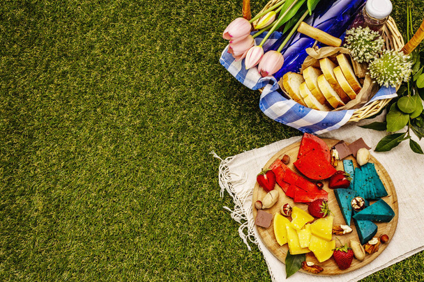 Picnic concept. Assorted multicolored hard Dutch cheeses. Blue and red pesto, aged gouda. Strawberry, chocolate, nuts, herbs, juice and wine. Wicker basket on green grass background, top view - Photo, Image
