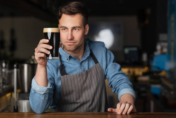 Bira ve barmen. Önlüklü yakışıklı adam bir bardak biraya bakıyor. - Fotoğraf, Görsel