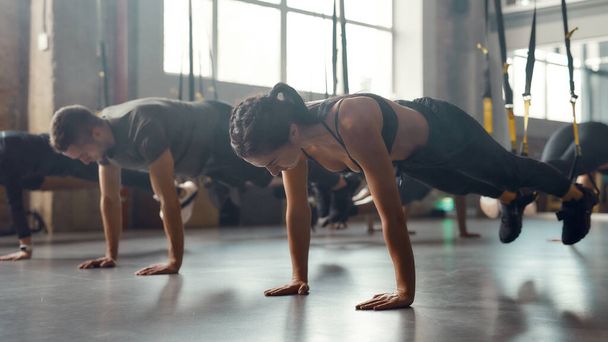 Keep balance. Athletic young woman stretching her body while doing fitness training exercises at industrial gym. Straps, group workout concept - 写真・画像