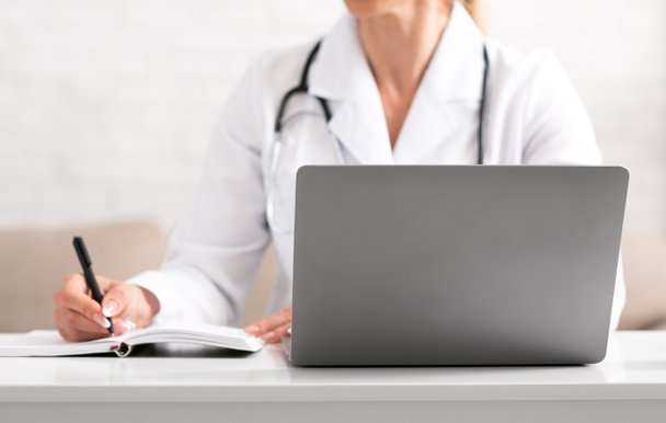 Online-Arztbesuche. Frau im weißen Mantel macht sich Notizen mit Stift und Notizbuch - Foto, Bild