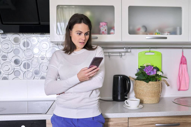 Giovane donna utilizzando smartphone, casa cucina interno sfondo - Foto, immagini