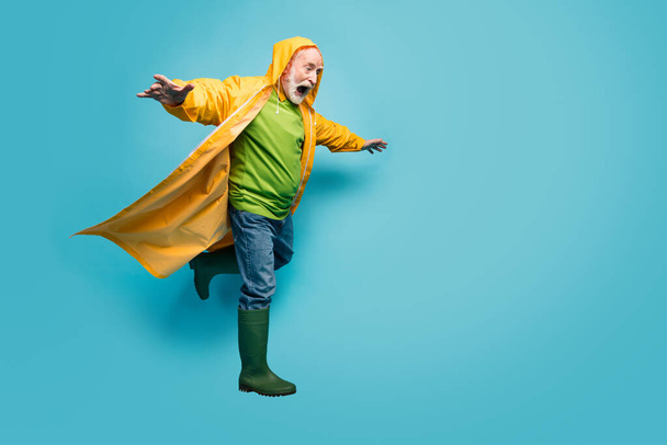 Full length photo of screaming grandpa amazed walk street puddles rainy weather slippery wet road wear jeans jumper gum boots yellow raincoat isolated blue color background - 写真・画像