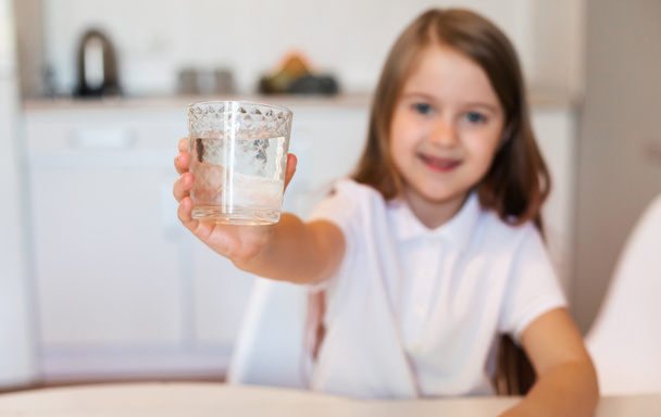 Κορίτσι που δείχνει ποτήρι νερό στην κάμερα που κάθεται στην κουζίνα - Φωτογραφία, εικόνα