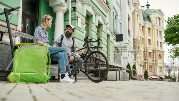 Dois correios alegres, jovem e mulher sentados no banco e conversando ao ar livre ao entregar alimentos e produtos, usando scooter e bicicleta
 - Foto, Imagem