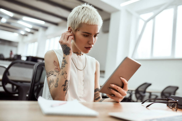 Skype call. Portrait of young cute blonde tattooed business woman putting on wireless earphones and using her touchpad while sitting in the modern office - 写真・画像