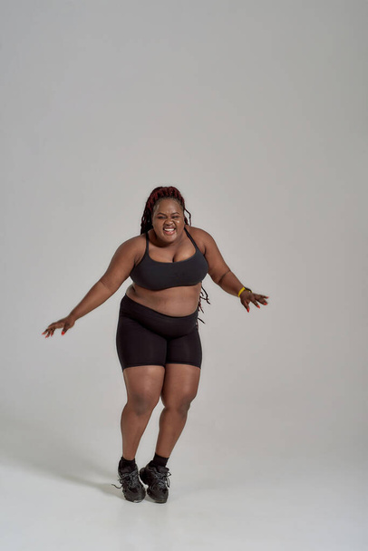 Fiercely love myself. Full length shot of plump, plus size african american woman in sportswear having fun, dancing in studio over grey background - Photo, image