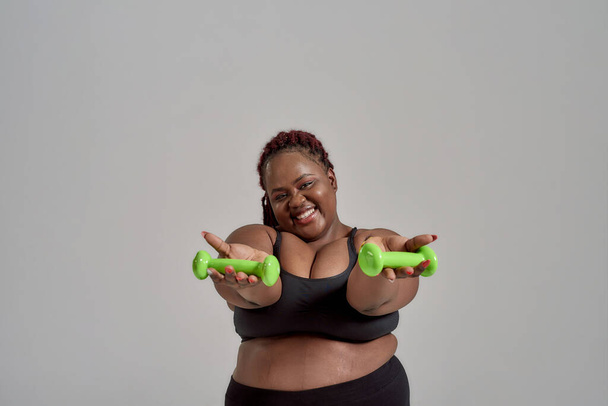 Plump, plus size african american woman in sportswear reaching her arms out, holding green dumbbells in studio over grey background - Photo, image
