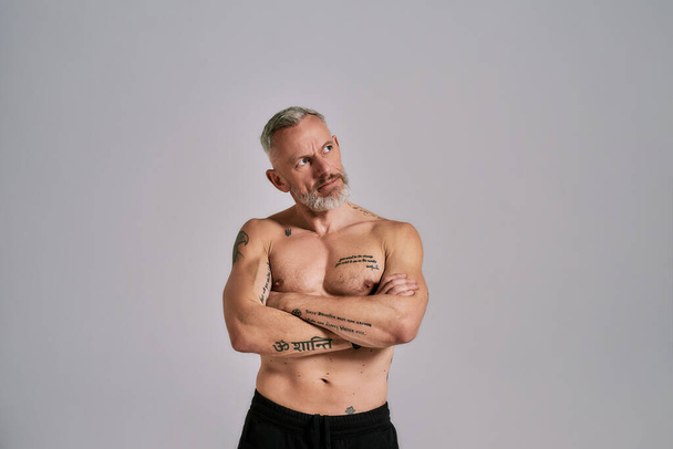 Half naked middle aged muscular man looking aside, showing his body, while posing in studio over grey background - Foto, Imagem