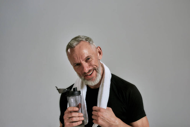 Middle aged muscular man in black t shirt smiling at camera holding bottle of water and towel around his neck, posing in studio over grey background - Photo, Image