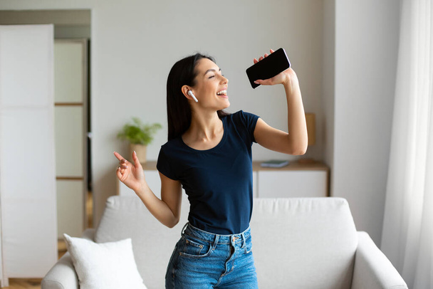 Девушка с наушниками и смартфоном поет песню, веселясь в помещении
 - Фото, изображение