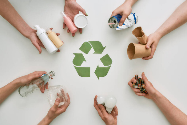 Πήγαινε πράσινο με την ανακύκλωση. Κάτοψη πολλών χεριών που κρατούν διαφορετικά απόβλητα, τύποι σκουπιδιών με πινακίδα ανακύκλωσης από χαρτί στο κέντρο πάνω από λευκό φόντο - Φωτογραφία, εικόνα