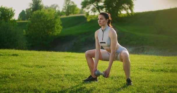 La vista dal lato delle donne accovaccia con il peso. Una donna si allena nel parco al mattino - Filmati, video