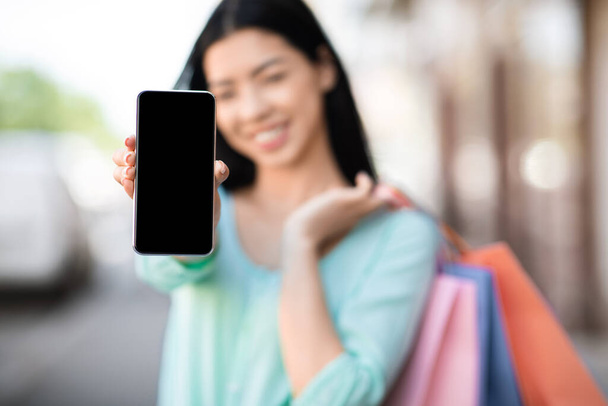 Aplicación de compras. Chica asiática con bolsas de comprador mostrando Smartphone con pantalla negra
 - Foto, Imagen
