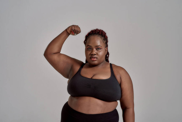My body. Plump, plus size african american woman in black sportswear looking at camera, showing strong arm in studio over grey background. Concept of sport, healthy lifestyle, body positive, equality - Photo, image