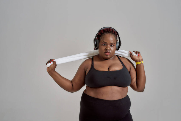 Getting the Best of My Body. Plump, plus size african american woman in sportswear and headphones holding towel, posing in studio over grey background - Фото, зображення