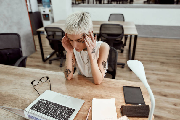 Hard working day. Young tired tattooed business woman keeping eyes closed, suffering from the headache while sitting at her working place in the modern office. Stress at work - 写真・画像