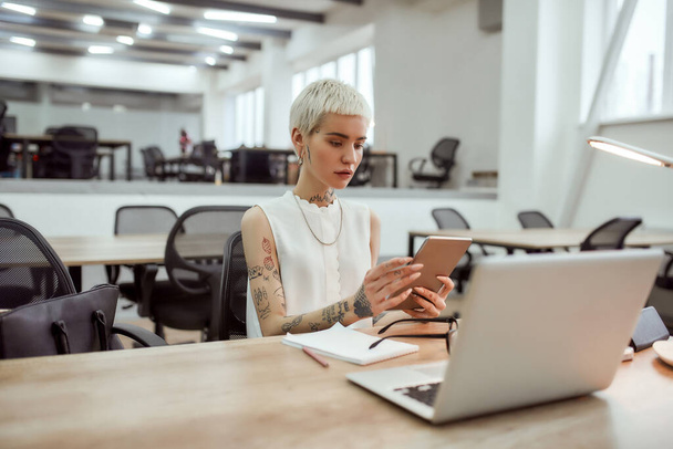 Full concentration. Portrait of young stylish blonde tattooed business woman with short haircut using touchpad while sitting at her working place in the modern office. Digital technologies at work - Photo, image