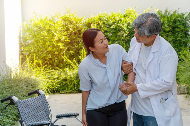 The doctor is helping to lift the patient out of the wheelchair while walking in the garden for physical therapy after the car accident. Concept of being by side and encouraging. - Photo, Image
