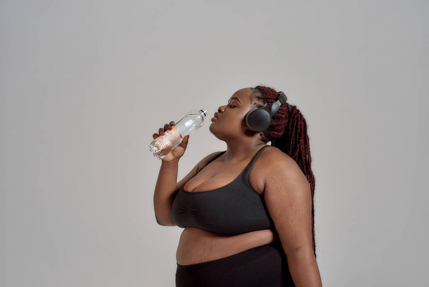 Care for your body. Plump, plus size african american woman in sportswear and headphones drinking water from the bottle, posing in studio over grey background - 写真・画像