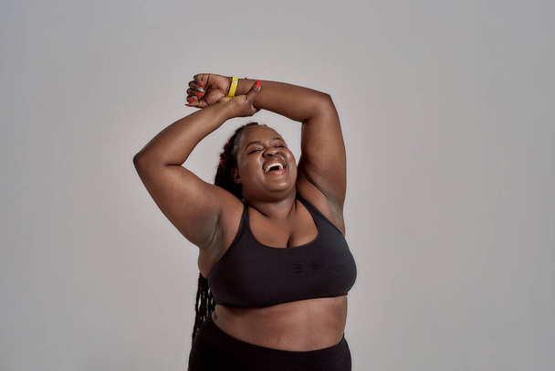 Love my body. Plump, plus size african american woman in sportswear laughing, lifting her arms up in studio over grey background. Concept of sport, healthy lifestyle, body positive, equality - Fotoğraf, Görsel