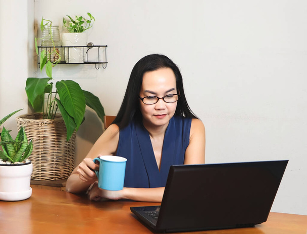 Aasian nainen istuu puinen pöytä sisätiloissa, tilalla kuppi kahvia ja katsot tietokoneen näytön pöydällä.Työskentely kotoa käsite. - Valokuva, kuva