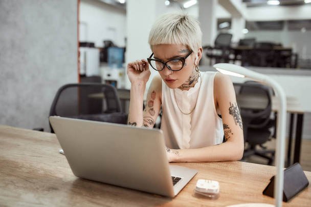 Ttrying to find a sulution. Thoughtful young blonde tattooed businesswoman in eyeglasses looking at the screen of laptop and thinking about something while sitting at her working place - Foto, immagini