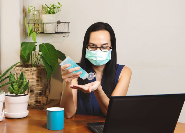 Asian woman wearing hygienic mask sitting at wooden table indoor with computer notebook ,washing her hands with alcohol sanitizer gel,working from home.protection from covid-19 concept. - Photo, Image
